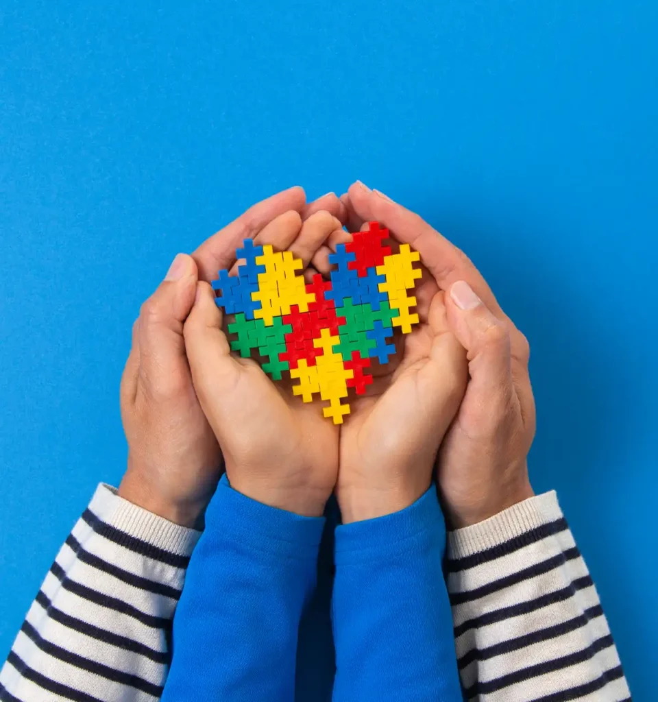 Hands holding a heart shape made of colorful puzzle pieces, symbolizing autism awareness, against a blue background.