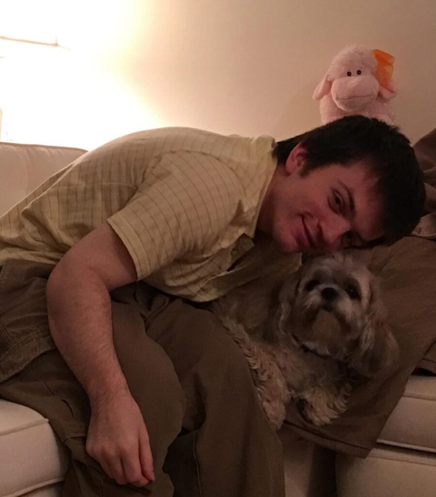 A man leans over on a couch, smiling while hugging a small dog with a toy on the backrest behind them.